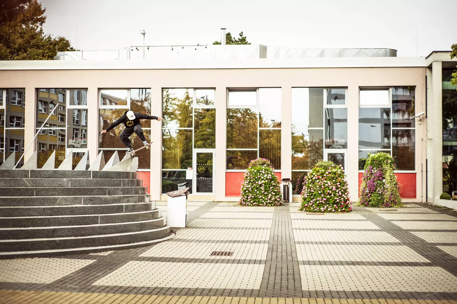 A skateboarder performing a trick on a staircase with uniquely shaped stone steps in front of a modern building, framed by potted flowers and reflective windows.