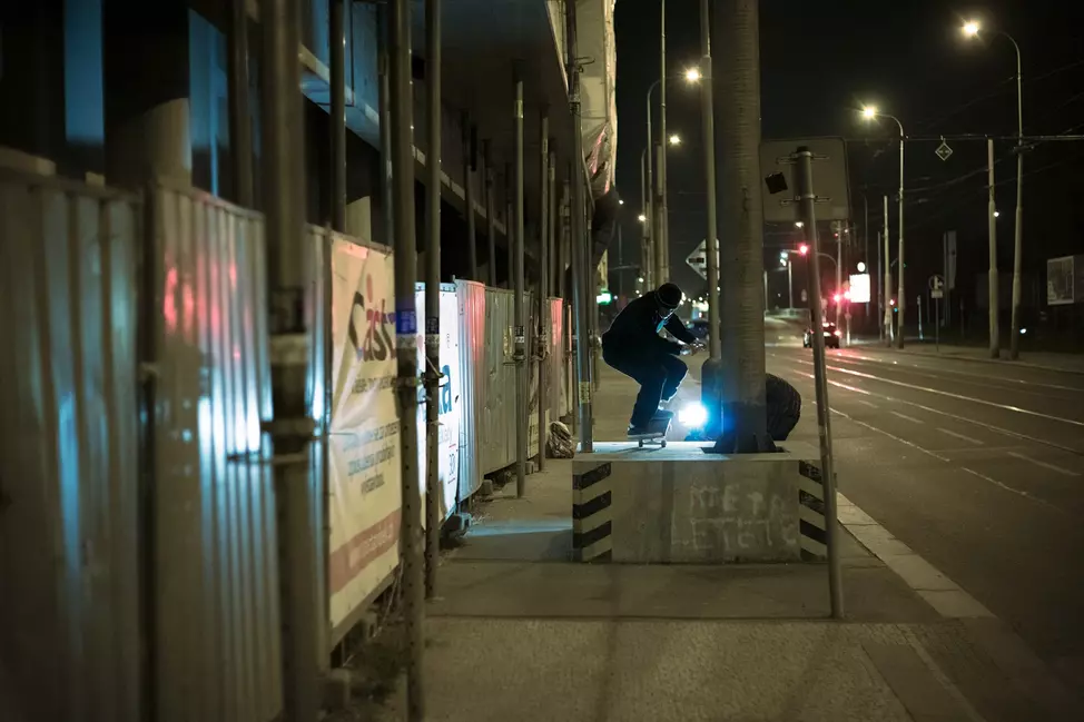 A nighttime street scene showing a person skating near a construction barrier illuminated by a bright light.